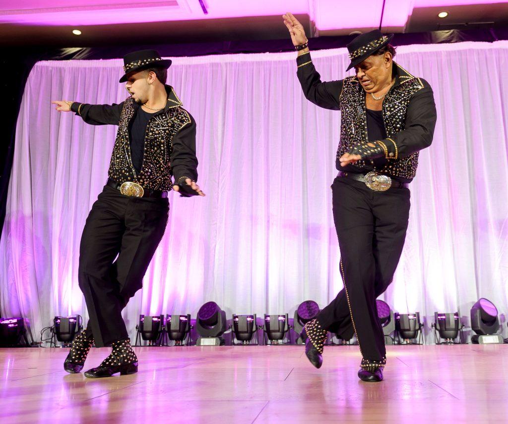 Dos bailarines de mambo con trajes negros y tachuelas doradas actuando en un escenario iluminado en rosa